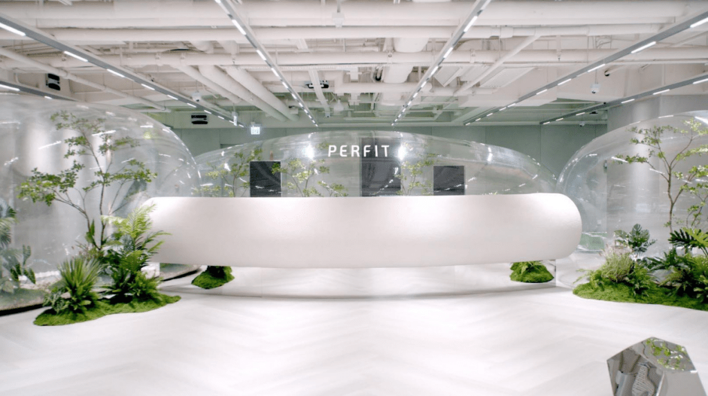 The bright, modern reception area of a perFIT gym featuring a sleek white desk and unique clear bubble enclosures filled with green plants.